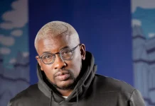 ‘Why women don’t treat men well,’ Do2dtun makes controversial claims Portrait of a man with short blond hair and round glasses wearing a black hoodie, standing in front of a blue, urban mural backdrop.