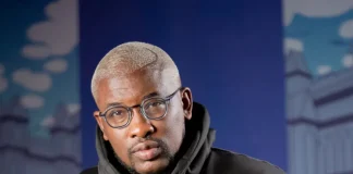 ‘Why women don’t treat men well,’ Do2dtun makes controversial claims Portrait of a man with short blond hair and round glasses wearing a black hoodie, standing in front of a blue, urban mural backdrop.