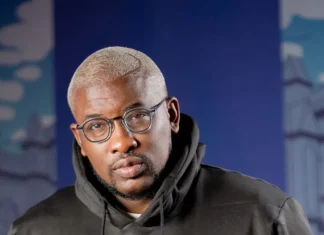 ‘Why women don’t treat men well,’ Do2dtun makes controversial claims Portrait of a man with short blond hair and round glasses wearing a black hoodie, standing in front of a blue, urban mural backdrop.