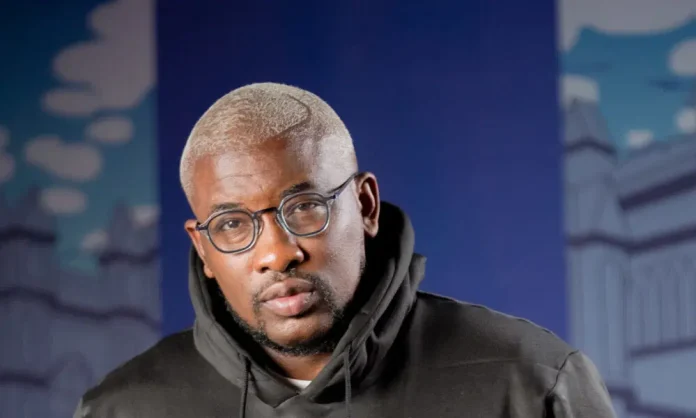 Portrait of a man with short blond hair and round glasses wearing a black hoodie, standing in front of a blue, urban mural backdrop.