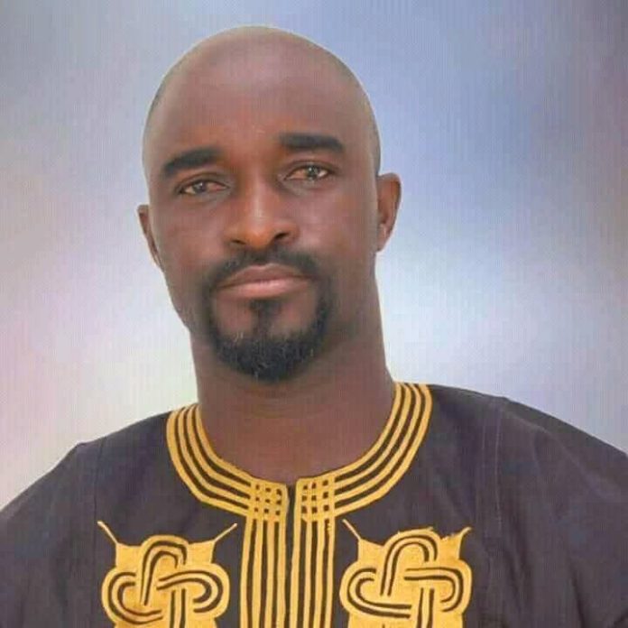 Portrait of a man with a goatee wearing a black traditional African outfit with gold embroidery, facing the camera.