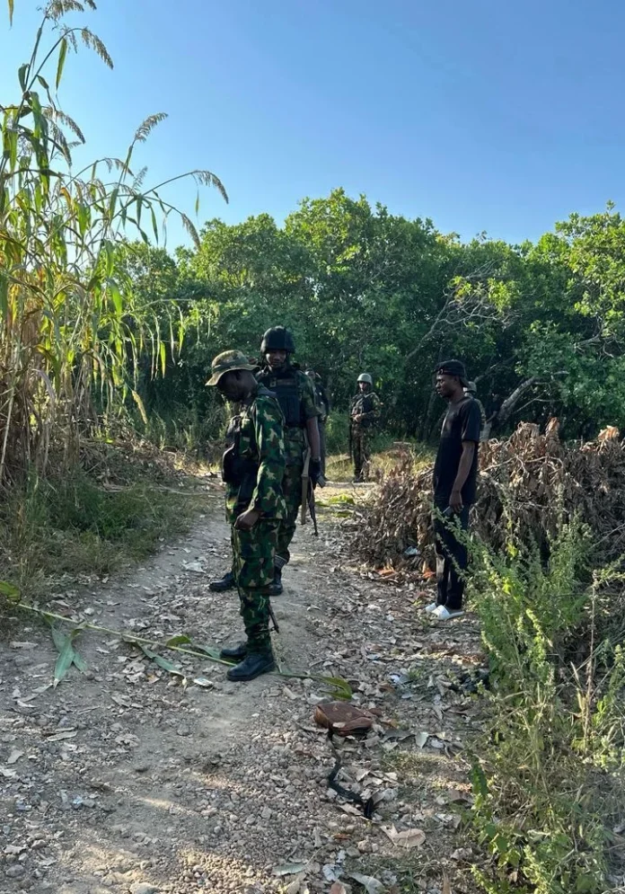Soldiers in camouflage gear with rifles stand on a rocky dirt path in a wooded area, a civilian man nearby wearing a black shirt and white sneakers.