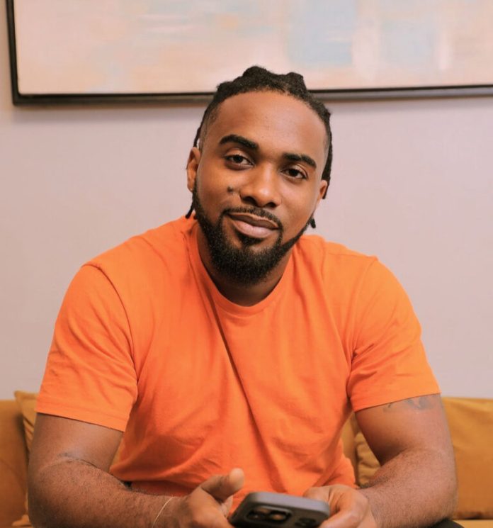 Smiling man in an orange T-shirt sitting on a couch, holding a smartphone toward the camera.