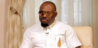 Nollywood is full of saboteurs – Jim Iyke Man in a white shirt with a gold pocket accessory and sunglasses sits on a beige chair for an interview, a mic clipped to his shirt.