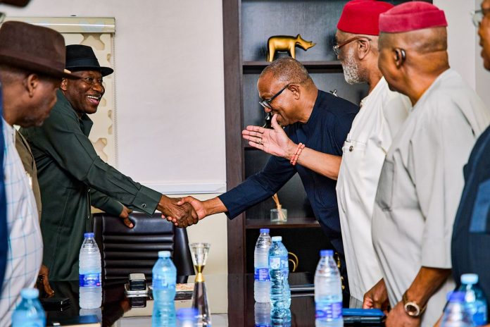 Two men in business attire shake hands across a conference table as others look on in a formal meeting setting.