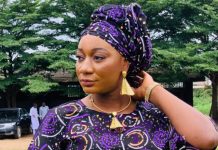 Leaked Video Scandal: Popular herbal vendor Sisi Alagbo breaks silence Woman in a purple patterned headwrap and dress, wearing gold jewelry, adjusting her headwrap outdoors with green trees in the background.