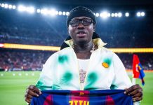 Barcelona recognises Nigerian entertainer Zlatan Ibile at Spotify Camp Nou Man on a stadium field holding a FC Barcelona jersey with the number 77, wearing a black head wrap and glasses, UNHCR branding visible at the bottom.