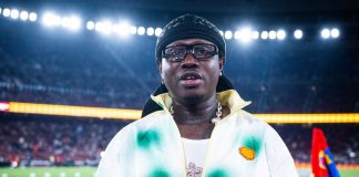 Barcelona recognises Nigerian entertainer Zlatan Ibile at Spotify Camp Nou Man on a stadium field holding a FC Barcelona jersey with the number 77, wearing a black head wrap and glasses, UNHCR branding visible at the bottom.