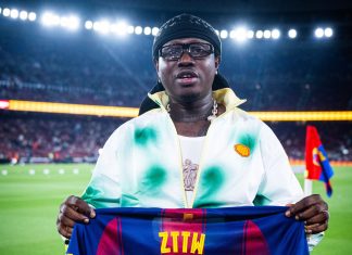Barcelona recognises Nigerian entertainer Zlatan Ibile at Spotify Camp Nou Man on a stadium field holding a FC Barcelona jersey with the number 77, wearing a black head wrap and glasses, UNHCR branding visible at the bottom.
