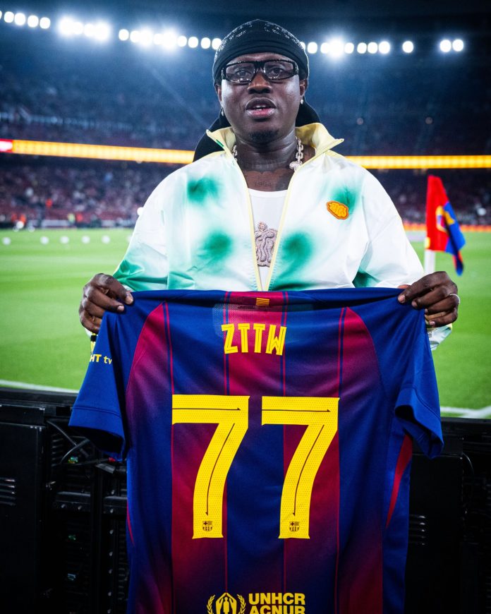 Man on a stadium field holding a FC Barcelona jersey with the number 77, wearing a black head wrap and glasses, UNHCR branding visible at the bottom.