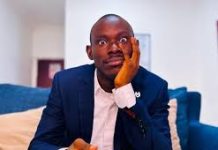 “Practice What You Preach?” — Gehgeh’s Sister Calls Him Out Over Relationship Advice Man in a navy suit sits on a blue sofa with a surprised, wide-eyed expression, resting his cheek on his hand and holding a small snack.