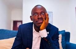 “Practice What You Preach?” — Gehgeh’s Sister Calls Him Out Over Relationship Advice Man in a navy suit sits on a blue sofa with a surprised, wide-eyed expression, resting his cheek on his hand and holding a small snack.