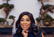 Mo Abudu warns public over fake video promising N700,000 daily earnings Woman in a navy dress smiling softly, resting her chin on her hand, wearing a silver watch and bracelet with a bonsai-filled backdrop behind her.