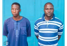 Two Abuja security guards bag life jail over serial rape of girl for 6 years Two men stand side by side in front of a bright blue door, wearing blue shirts (left in a plain tunic, right in a striped polo).