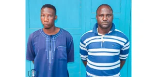 Two Abuja security guards bag life jail over serial rape of girl for 6 years Two men stand side by side in front of a bright blue door, wearing blue shirts (left in a plain tunic, right in a striped polo).