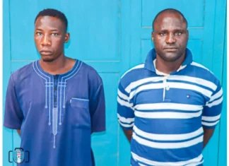 Two Abuja security guards bag life jail over serial rape of girl for 6 years Two men stand side by side in front of a bright blue door, wearing blue shirts (left in a plain tunic, right in a striped polo).
