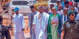 Rep seat: Yusuf Buhari gets automatic APC ticket Group of men in traditional Nigerian attire walking together, escorted by a security officer in front, while a photographer films on a sunny street.