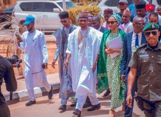 Rep seat: Yusuf Buhari gets automatic APC ticket Group of men in traditional Nigerian attire walking together, escorted by a security officer in front, while a photographer films on a sunny street.