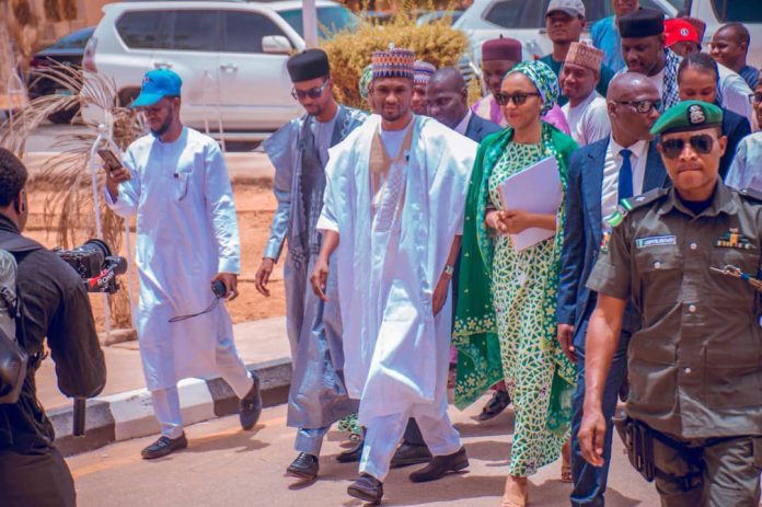 Group of men in traditional Nigerian attire walking together, escorted by a security officer in front, while a photographer films on a sunny street.