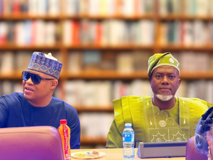 Two men in colorful traditional outfits sit at a table in a book-lined room; the left man wears blue with sunglasses and a patterned cap, the right wears green with a head wrap.