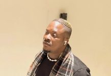 What were you expecting? — Jaywon questions Ilebaye’s father Man wearing a black t‑shirt with a plaid scarf, white shell necklace, and AirPods, looking to the side indoors.