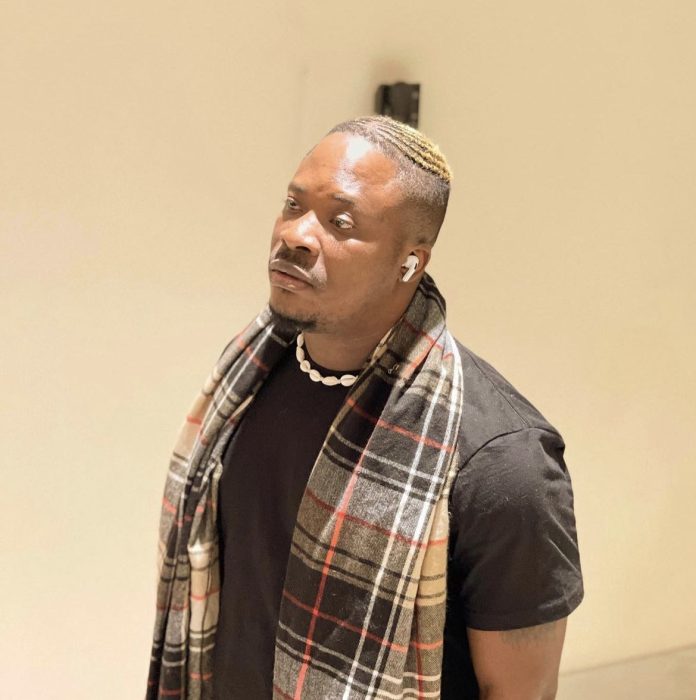 Man wearing a black t‑shirt with a plaid scarf, white shell necklace, and AirPods, looking to the side indoors.