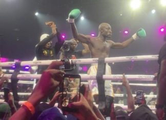 E-Money rewards Carter Efe with cash gift after defeating Portable Boxer with green gloves raises arms in victory inside a packed boxing ring as fans in the crowd film and cheer under bright stage lights.