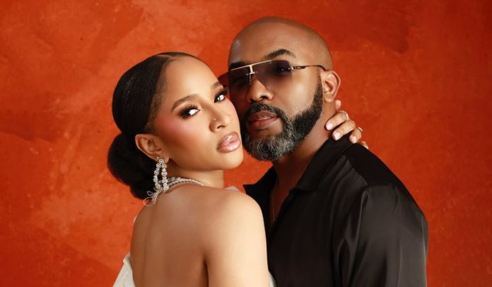 Couple posing closely against an orange textured backdrop; woman wears a white strapless dress and crystal chandelier earrings, man wears sunglasses and a black shirt.