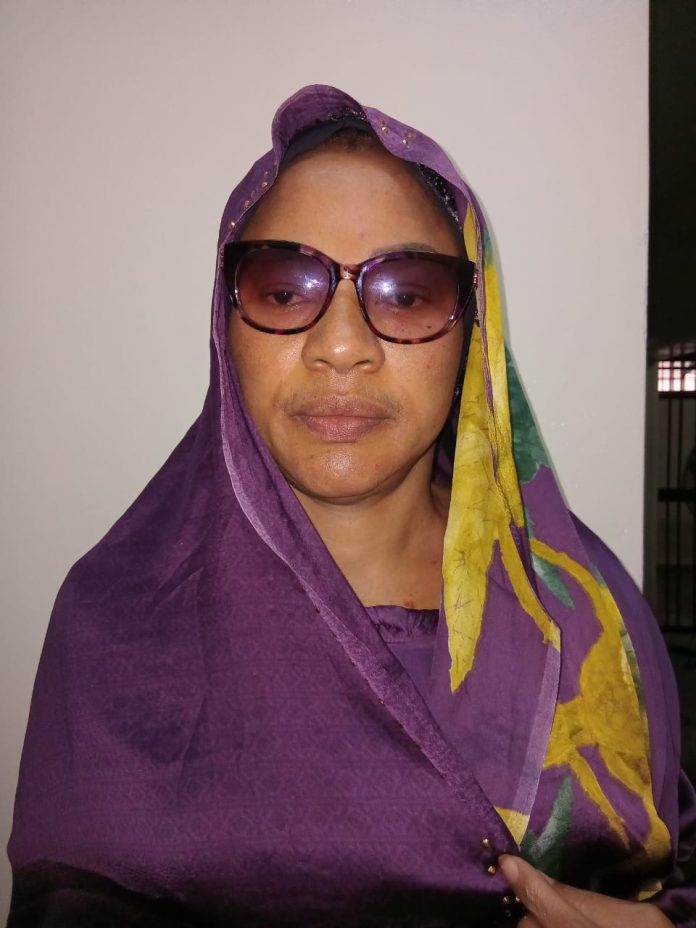 Portrait of a woman wearing a purple hijab and a yellow-green patterned scarf, sunglasses, and a neutral expression, facing the camera.