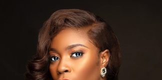 YouTube was never designed for films – Bolaji Ogunmola speaks on filmmaking Portrait of a woman with wavy brown hair, bold makeup (red lips, subtle eye makeup), wearing a teal V-neck dress and a gold earring against a dark backdrop.