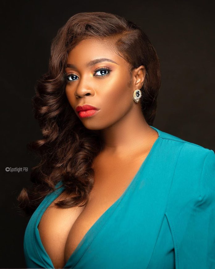 Portrait of a woman with wavy brown hair, bold makeup (red lips, subtle eye makeup), wearing a teal V-neck dress and a gold earring against a dark backdrop.