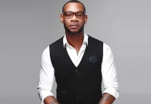 D’Prince decries monetisation of cyberbullying in entertainment industry Portrait of a stylish man in a white shirt with a black vest, wearing round glasses, with a tattoo visible on his forearm, against a gray studio backdrop.