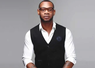 D’Prince decries monetisation of cyberbullying in entertainment industry Portrait of a stylish man in a white shirt with a black vest, wearing round glasses, with a tattoo visible on his forearm, against a gray studio backdrop.
