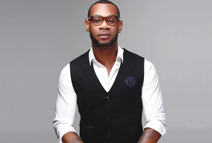 Portrait of a stylish man in a white shirt with a black vest, wearing round glasses, with a tattoo visible on his forearm, against a gray studio backdrop.