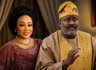 “I Stepped Down for Desmond Elliot in 2023” — Barakat Bakare Speaks on Surulere Political Tension Smiling woman in a black gele and burgundy traditional dress with pearl jewelry, beside a man in gold-embroidered agbada in a warmly lit room.