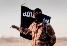 How we kiiled ISIS leader in collaboration with US forces – Nigerian military Masked militant in desert camouflage points to the left with a black flag bearing white Arabic script behind him.