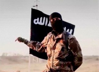 How we kiiled ISIS leader in collaboration with US forces – Nigerian military Masked militant in desert camouflage points to the left with a black flag bearing white Arabic script behind him.