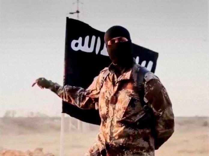 FB_IMG_1778925683635 1 Masked militant in desert camouflage points to the left with a black flag bearing white Arabic script behind him.