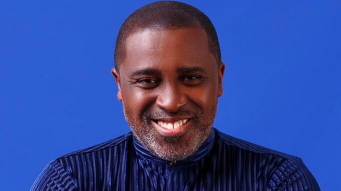 Head-and-shoulders portrait of a smiling Black man in a dark striped shirt against a blue backdrop.