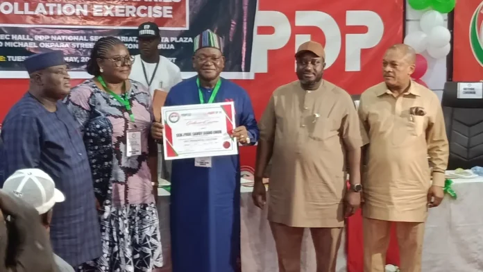 Six people stand on a stage during an awards event; a man in blue traditional attire holds a framed certificate as others pose nearby.