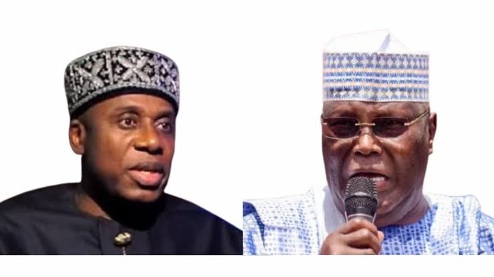 Two men in traditional Nigerian attire; left man wears a patterned black cap, right man wears a light blue patterned cap and speaks into a microphone.