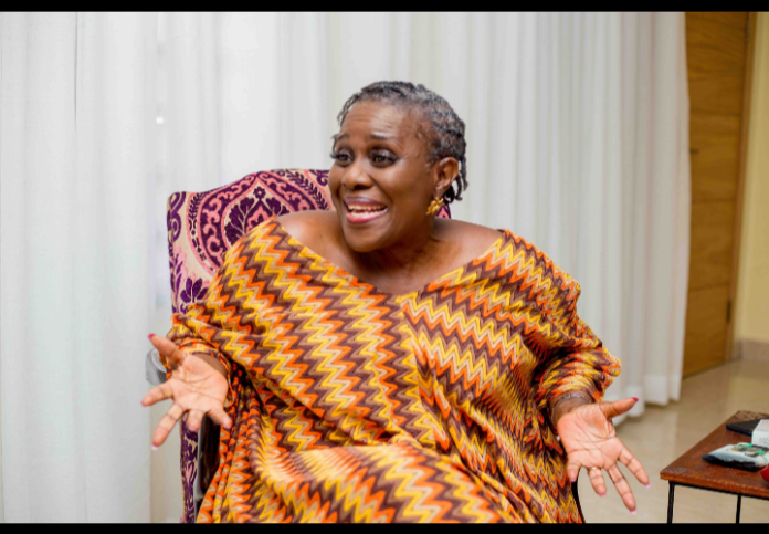 A smiling woman in a colorful zigzag dress sits on a patterned chair, gesturing with open hands as she speaks.
