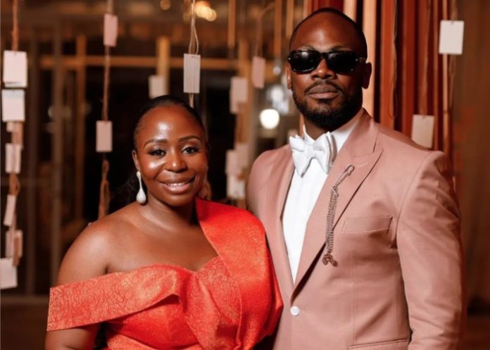 Couple posing together at a formal event; woman in a red off-shoulder dress, man in a light pink suit and sunglasses.