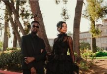 Mr Eazi, Otedola’s daughter Temi, celebrate first wedding anniversary Two individuals in black formal outfits posing on a sunny street framed by trees; man wears a suit with sunglasses, woman in a voluminous black gown.