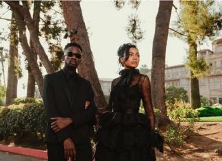 Mr Eazi, Otedola’s daughter Temi, celebrate first wedding anniversary Two individuals in black formal outfits posing on a sunny street framed by trees; man wears a suit with sunglasses, woman in a voluminous black gown.