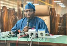 INEC breaks silence on plot to deregister ADC ahead of 2027 Elections Man in blue traditional attire speaking into a handheld mic at a press conference, with several microphones on a glass table in front of him.