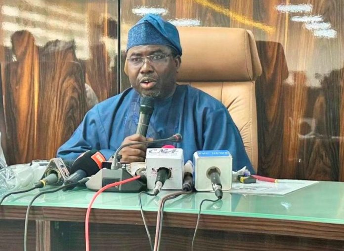Man in blue traditional attire speaking into a handheld mic at a press conference, with several microphones on a glass table in front of him.
