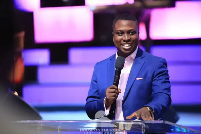 Smiling man in a blue suit speaks into a microphone at a podium on a lit purple stage.