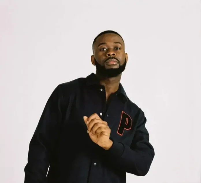 Sarx-e1694910045808 2 Portrait of a man with a beard wearing a black jacket with a red P patch, looking at the camera.