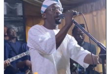 Malaika Warms Hearts After Sweet Gesture to Iya Rainbow at Event (Video) Male singer in white traditional outfit performs on stage with a live band behind; FASIWAJU TV logo overlay.